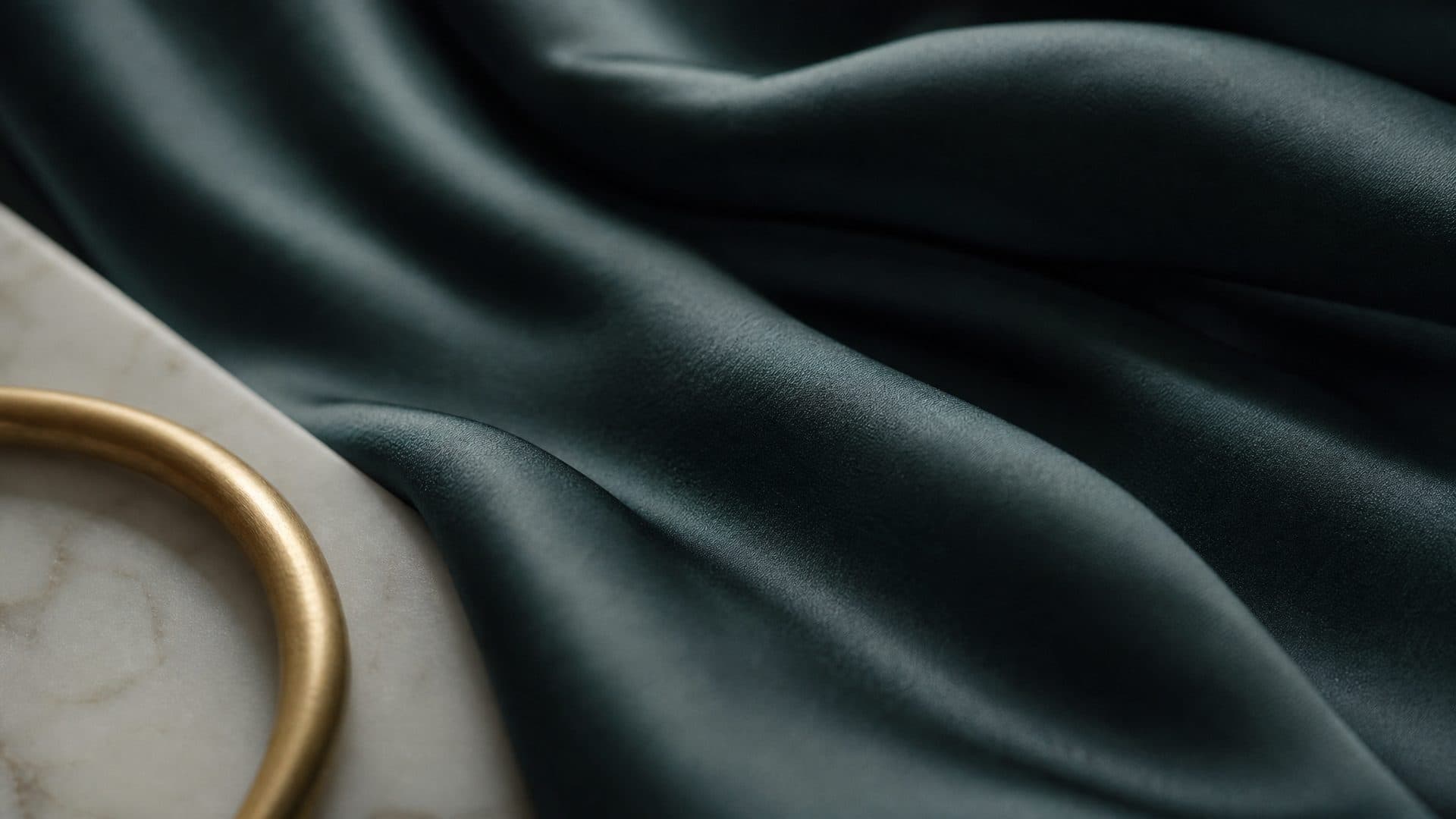 Deep teal satin fabric in soft folds with a brass ring resting on it, the light sliding along the folds.