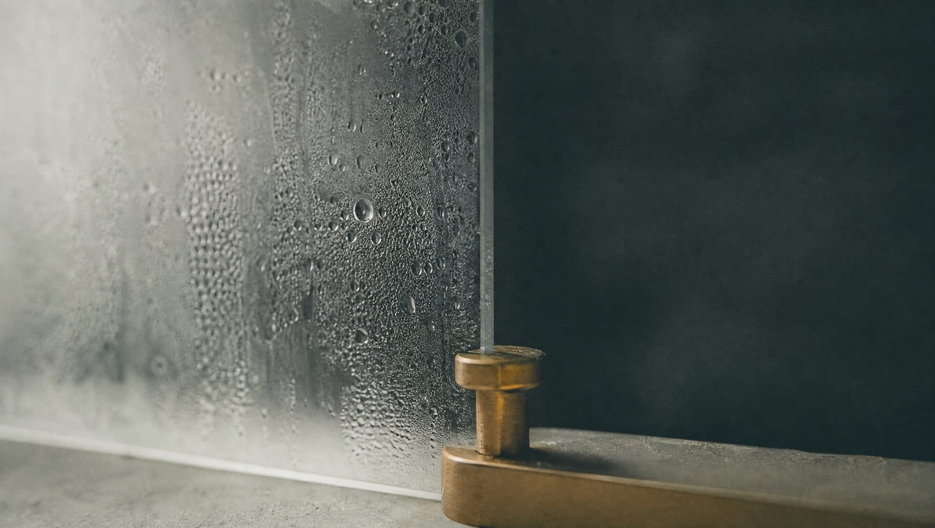 Condensation and fine droplets on a cool pane of glass, with a deep teal blur and a brass fitting behind it.