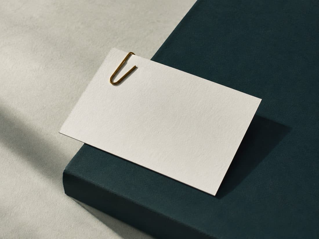 A single off-white card lying slightly askew on a deep teal surface, held at its corner by a small brass paperclip, with a hard shadow beneath it.