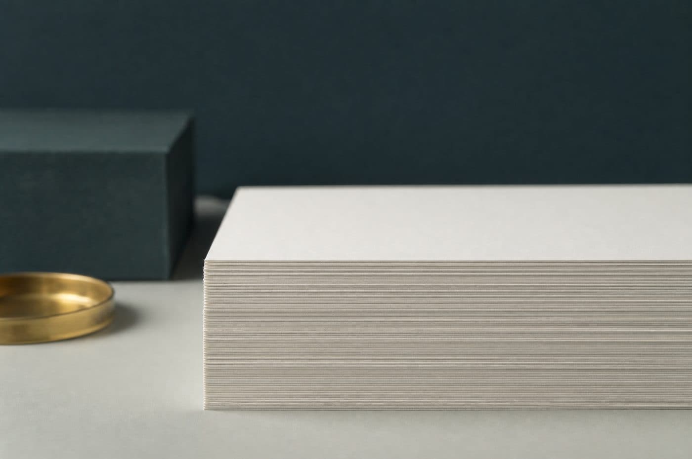 A stack of off-white card cut perfectly level across the front edge, the individual sheet edges reading as fine horizontal lines, beside a shallow brass dish.