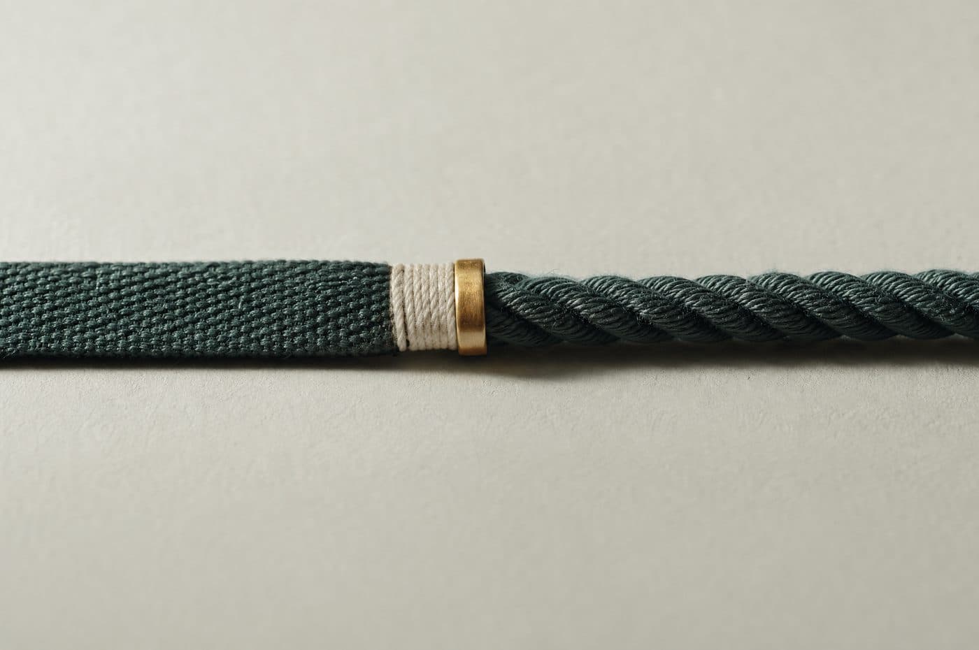 A cord that begins as a flat woven band and becomes a round twisted rope along its length, joined by a brass collar, on pale paper.