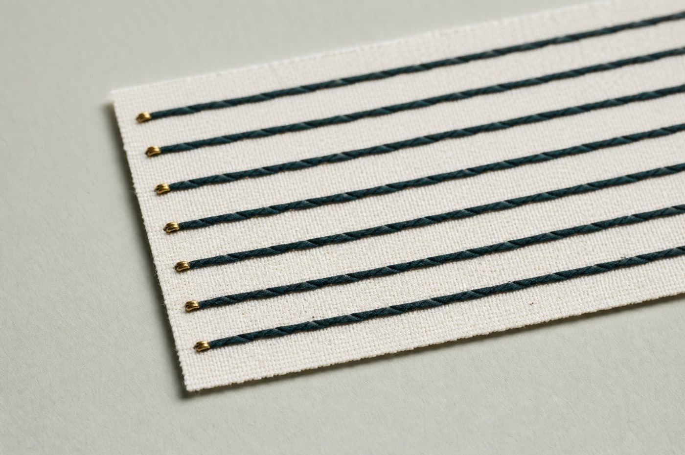A stiff off-white card ruled with six evenly spaced deep teal lines, each one pinned at the edge with a small brass stud.